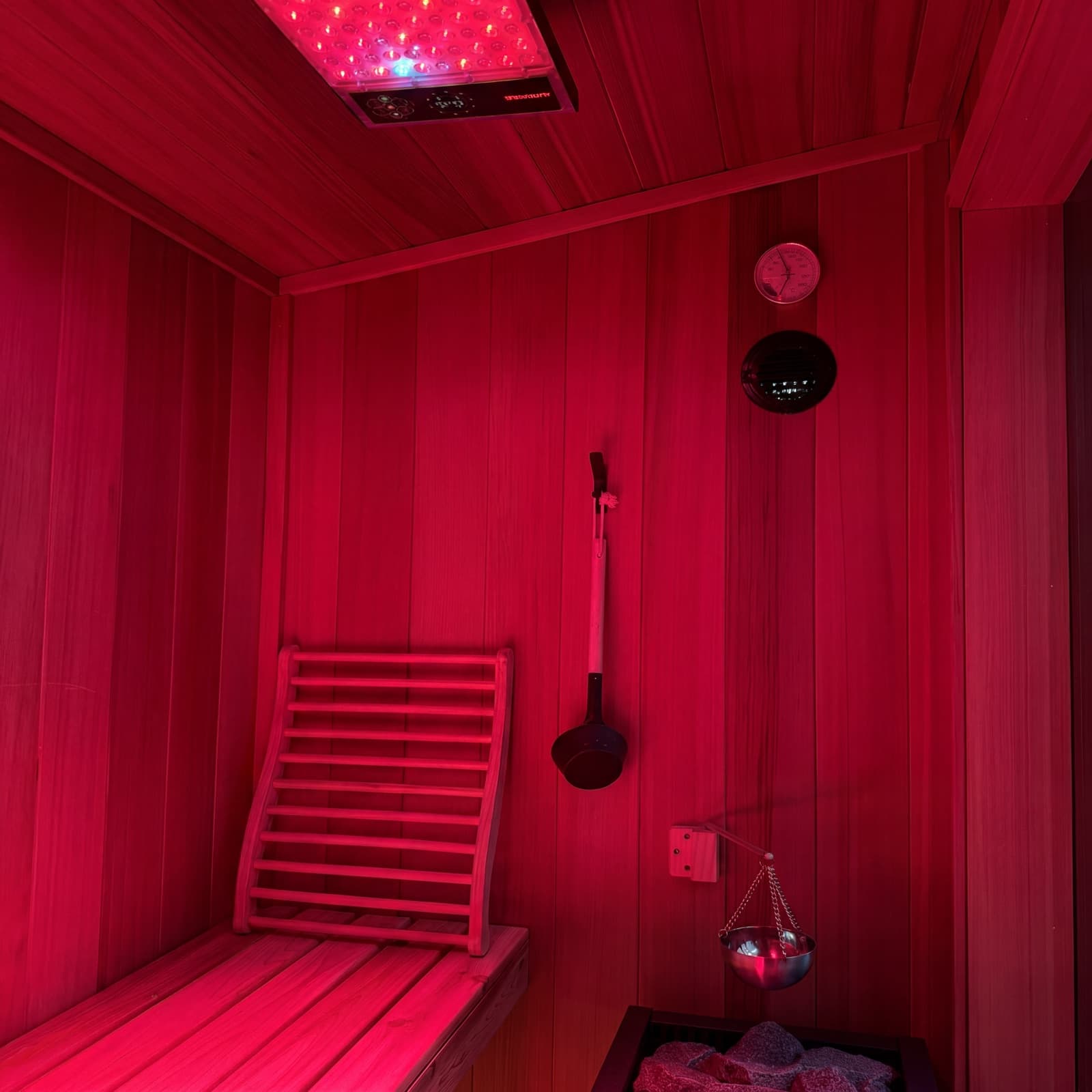 Sauna interior bathed in red light from the overhead therapy panel — cedar bench with backrest, ladle, thermometer, and sand-timer on the wall